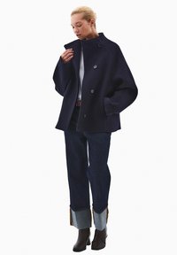 Przesadnie duży granatowy wełniany płaszcz z wysokim kołnierzem, zapięciem na guziki i opadłymi ramionami. Połączony z ciemnymi dżinsami z mankietami i brązowymi botkami za kostkę.