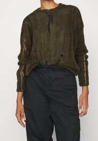 Olivgrön stickad tröja med stora hål och slitna texturer. Långa ärmar med synliga ribbade detaljer. Kombinerad med svarta baggy-byxor.