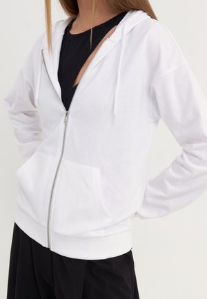 Vrouw die een witte hoodie met rits draagt over een zwart shirt, met haar handen op haar heupen tegen een effen achtergrond.