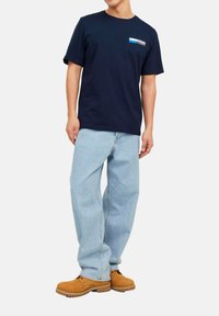 Marineblå bomulds t-shirt med korte ærmer, der har et lille logo i hvidt og blåt. Kombineret med lyseblå denimjeans og brune arbejdssko.