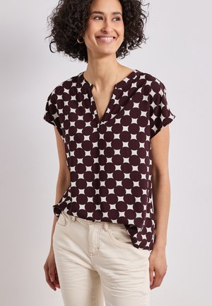 Femme aux cheveux bouclés souriante, portant un chemisier à manches courtes bordeaux à motifs ronds blancs et un pantalon beige clair.