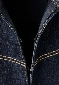 Tissu en denim bleu foncé avec double surpiqûre jaune et fermetures métalliques à crochets et œil partiellement fermées sur une ouverture de vêtement.