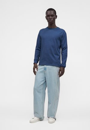 Homme portant une chemise bleue à manches longues, un jean large bleu clair et des baskets blanches, debout devant un fond blanc uni.