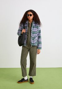 Giacca in fleece a motivi a quadri verdi e viola con zip e due tasche frontali, indossata sopra una tuta verde oliva e scarpe da ginnastica nere.