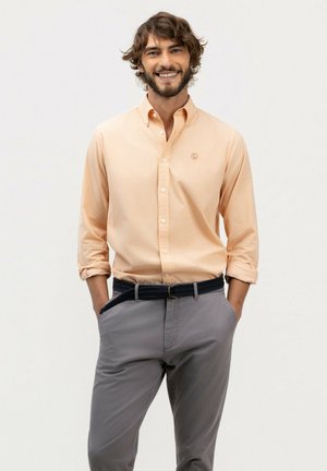 Hombre sonriente con cabello rizado que lleva una camisa de botones color melocotón claro y pantalones grises con las manos en los bolsillos frente a un fondo liso.