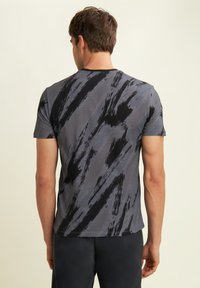 Hombre vistiendo una camiseta de manga corta gris y negra con un patrón abstracto, visto desde atrás, de pie contra un fondo liso.