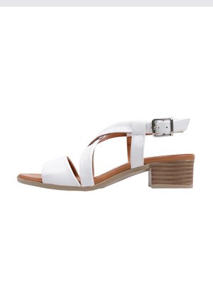 Sandalia de cuero blanca con correas cruzadas, hebilla ajustable en el tobillo y un tacón bajo de bloque marrón texturizado. Suela suave con ligero agarre.