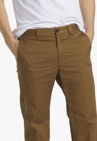 Braune Baumwollhosen mit glatter Textur, geradem Schnitt, Gürtelschlaufen und einem Knopfverschluss. Zwei seitliche Taschen sichtbar.