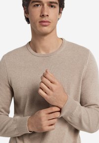 Ung man som justerar ärmen på en beige stickad tröja, stående mot en enkel vit bakgrund.