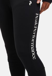 Schwarze Performance-Leggings aus glattem, dehnbarem Material mit weißem "Peak Performance"-Logo an der Seite. Akzentnähte sichtbar.