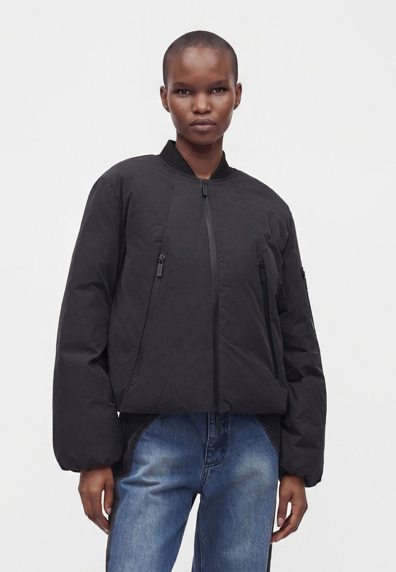 Άτομο που φοράει μαύρο bomber jacket με φερμουάρ και δύο εμπρόσθιες τσέπες με φερμουάρ, καθώς και μπλε τζιν, στέκεται μπροστά σε απλό ανοιχτό φόντο.