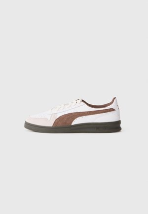 Biały sneaker z brązowym zamszowym paskiem, ciemną podeszwą i sznurowanym fasonem, pokazany z profilu na jasnym, jednolitym tle.
