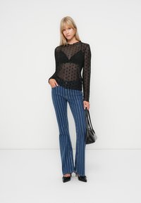 Zwarte doorzichtige top met lange mouwen en stippen, gecombineerd met blauwe gestreepte flared jeans en zwarte hakken, terwijl er een klein zwart tasje vastgehouden wordt.