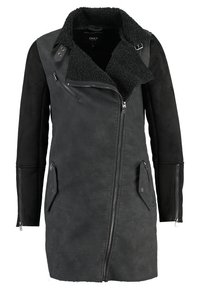 Musta tekonahkatakki, jossa on teksturoitu kaulus, epäsymmetrinen vetoketju ja kaksi etutaskua; yhdistää matta- ja hieman kiiltäviä materiaaleja.