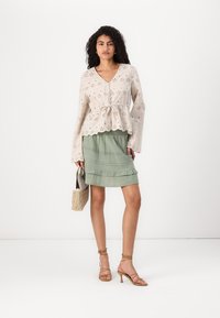 Light beige blouse with eyelet pattern and tie detail, paired with a green ruffled skirt. Tan strappy heels and a woven bag complete the look.