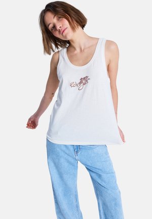 Jeune femme inclinant la tête, portant un haut blanc sans manches avec un logo floral "Roxy" et un jean bleu clair ample, sur un fond blanc.