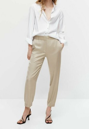 Woman wearing beige high-waist tapered pants, white button-down shirt with rolled sleeves, and black strappy heeled sandals, standing with hands in pockets.