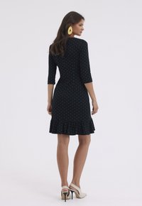 Czarna sukienka w kropki z rękawami o długości trzech czwartych, dopasowanym fasonie w górnej części, rozkloszowanym hemie z falbanką i gładką teksturą materiału. Noszona na wysokich obcasach.