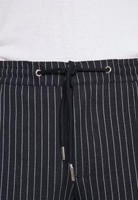 Spodnie w czarno-białe paski z sznurkiem w pasie, zakończonymi metalowymi, srebrnymi końcówkami, wykonane z fakturowanej tkaniny.