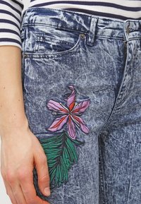 Jeans dżinsowe w wyblakłym niebieskim kolorze, z różowym i zielonym haftem kwiatowym na lewej udzie. Klasyczny pięciokieszeniowy fason.