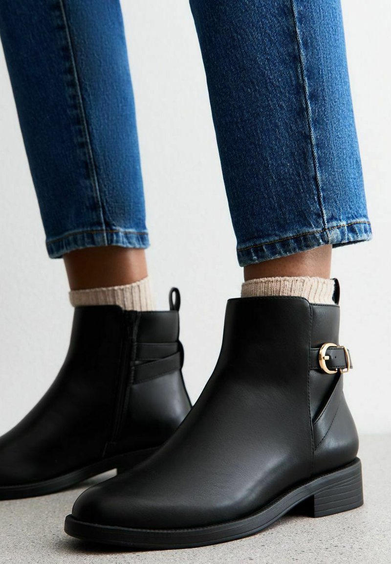 Botines de cuero negro con una textura suave, presentan un detalle de hebilla dorada y un tacón bajo apilado. Se combinan con jeans azules y calcetines beige.