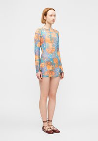 Νεαρό άτομο όρθιο, φοράει μια μακριά, πολύχρωμη διαφανή μίνι φόρεμα με αφηρημένα πορτοκαλί και μπλε σχέδια και καφέ παπούτσια με κορδόνια.