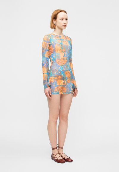 Νεαρό άτομο όρθιο, φοράει μια μακριά, πολύχρωμη διαφανή μίνι φόρεμα με αφηρημένα πορτοκαλί και μπλε σχέδια και καφέ παπούτσια με κορδόνια.