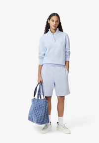 Felpa azzurra con zip e pantaloni corti coordinati. La modella tiene una borsa a spalla blu con un motivo. Sneakers bianche con dettagli verdi.