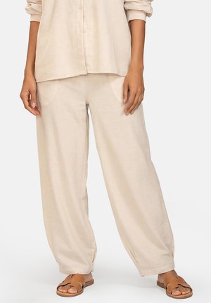 Luźne beżowe spodnie z przednimi kieszeniami, zestawione z pasującą koszulą na guziki i brązowymi sandałami z odkrytymi palcami na stojącej osobie.