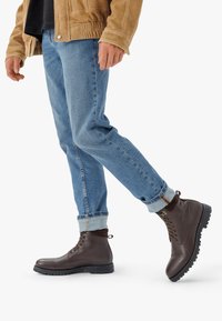 Bărci de piele maronie până la gleznă, cu șireturi, talpă din cauciuc și o buclă la călcâi, purtate cu blugi denim răsuciți și o jachetă de culoare deschisă.