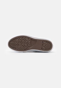 Hnědá gumová podrážka boty s texturovaným povrchem, obsahující geometrické vzory a bílý okraj. Branding je viditelný v oblasti paty.