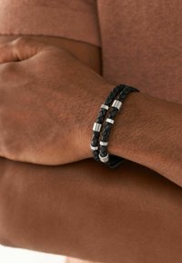 Schwarzes geflochtenes Lederarmband mit silbernen Metallakzenten, das glatte und strukturierte Abschnitte aufweist, getragen am Handgelenk vor einem braunen Hintergrund.