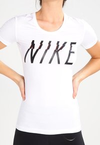 Camiseta blanca de manga corta con un prominente logo negro de "NIKE" con acentos rojos. Hecha de una tela ligera, tiene un diseño ajustado.