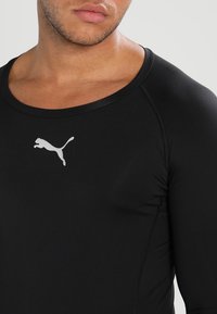 Sort atletisk langærmet skjorte med rund halsudskæring og glat, strækbart stof; har et hvidt Puma-logo på forsiden.