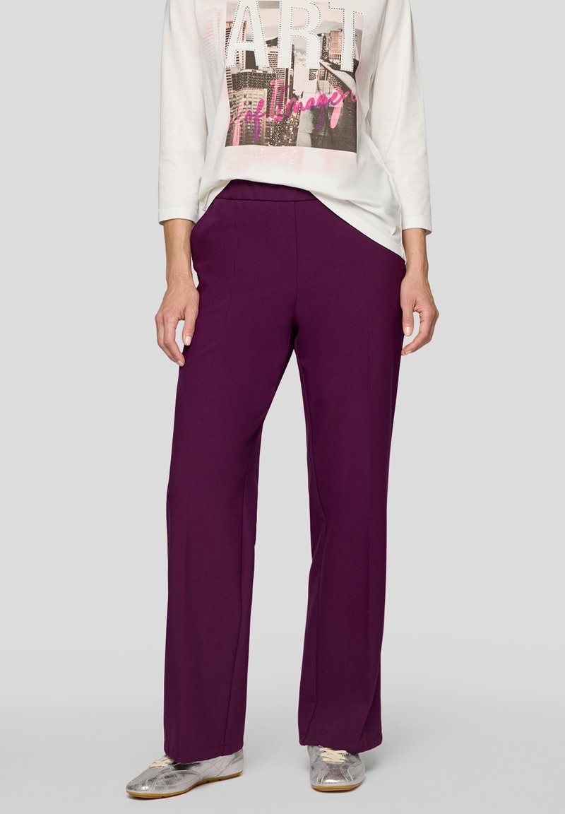 Purple wide-leg trousers in a soft fabric, paired with a white long-sleeve top featuring a graphic print. Silver sneakers complete the outfit.