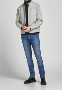 Man som bär en ljusgrå zip-up jacka, svart rullkrage, blå jeans och svarta snörskor, står mot en enkel ljus bakgrund.