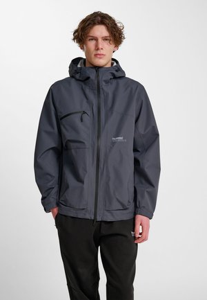 Ung mand iført en mørkegrå hættetrøje med lynlåslommer og sorte bukser, stående foran en ensfarvet, lys baggrund.