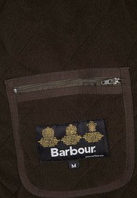 Brązowy materiał pikowany z kieszenią na zamek i metką z złotymi koronami oraz napisem "Barbour". Etykieta rozmiaru "M".