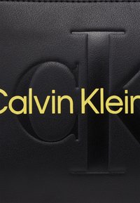 Černá kožená taška s vyra�ženým logem. Text "Calvin Klein" je v tučném žlutém písmu, které kontrastuje s texturovaným povrchem.