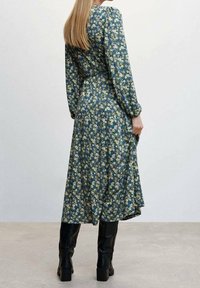 Kleid mit Blumenmuster auf grünem Hintergrund, langen Ärmeln und taillierter Taille. Getragen mit knielangen schwarzen Stiefeln.