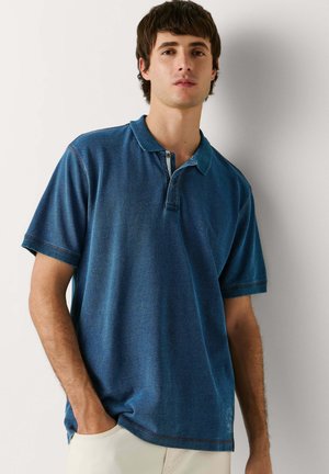 Jeune homme portant un polo bleu à manches courtes et un pantalon beige clair, debout avec une main dans la poche, devant un fond uni.