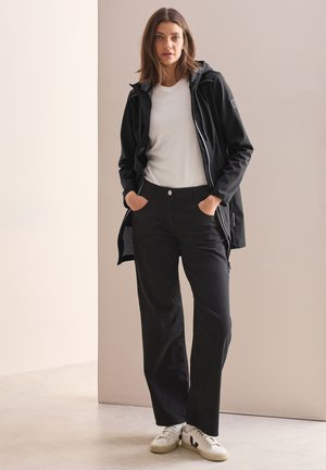 Giacca softshell nera con cappuccio, t-shirt bianca sotto e pantaloni neri a gamba larga. Sneakers bianche con dettagli neri. Design minimalista.