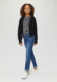 Schwarze Strickjacke über einem floral gemusterten Hemd, kombiniert mit blauen Jeans und weißen Sneakers. Lockige Haare, lässige Haltung, neutraler Hintergrund.