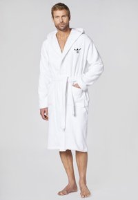 Weißer Frottee-Bademantel mit Kapuze, vorderem Bindegürtel und zwei seitlichen Taschen. Mit schwarzem Logo auf der Brust. Weicher, strukturierter Stoff.