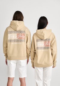 Beige huva sweatshirt med stor grafisk text på baksidan, inklusive platsnamn och "Sedan 1985 New York". Tillverkad av bomull, avslappnad passform.