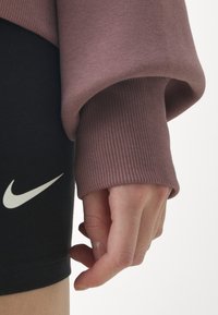 Pitkähihainen luumuinen pusero, jossa on resorit hihansuissa, yhdistetty mustiin shortseihin, joissa on valkoinen Nike-logo, sileä kangas ja istuva malli.