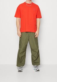 T-shirt en coton rouge à col rond associé à un pantalon cargo vert olive ample avec des poches latérales. Porté avec des baskets métalliques argentées.