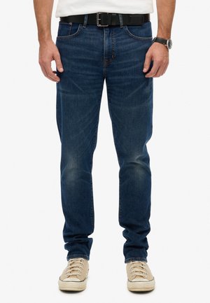 Man in een donkerblauwe spijkerbroek, witte sneakers, een zwarte leren riem, een wit shirt en een zwart horloge, staande tegen een witte achtergrond.