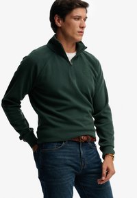 Ciemnozielona polarowa bluza rozpinana na zamek z prążkowaną fakturą i rękawami typu raglan, z logo na piersi, noszona z niebieskimi dżinsami.