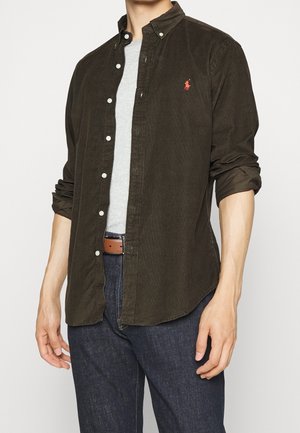 Mand iført mørkebrun kordfløjls-skjorte med knapper over en lysegrå t-shirt, blå jeans og brun læderbælte, ærmerne rullet op.